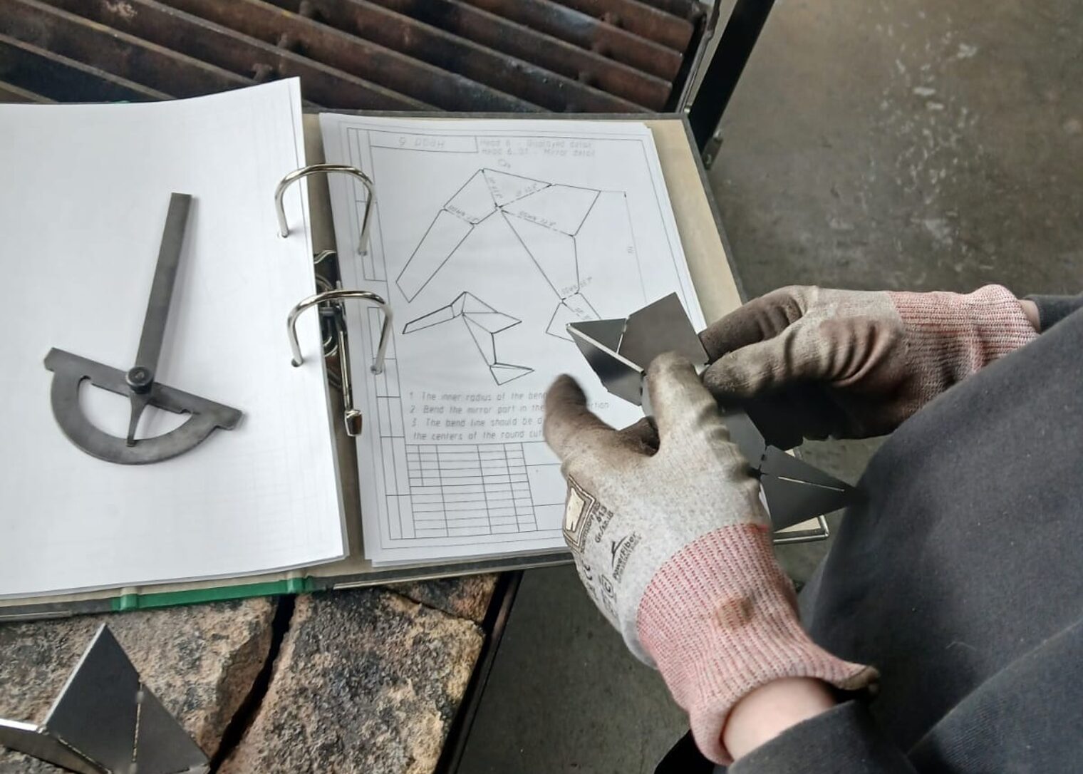 Hände mit Arbeitshandschuhen halten zugeschnittene Metallteile über einer technischen Zeichnung. Daneben liegen Werkzeuge und ein Ordner mit Bauplan für ein Metallprojekt.