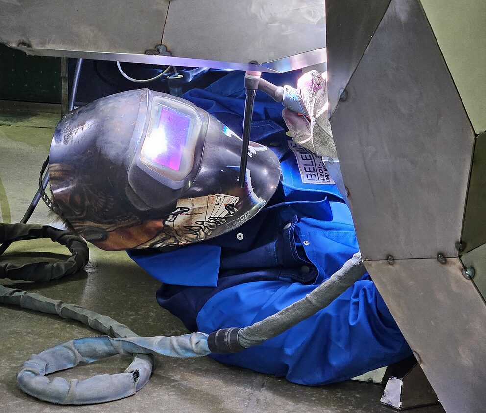 Schweißer in blauem Schutzanzug und Schweißhelm schweißt in einer engen Überkopfposition an der Metall-Gorilla-Skulptur beim DVS-Schweißevent der BBT Tuttlingen.